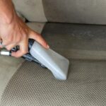 Hands using a vacuum nozzle to deeply clean a fabric car seat, removing dirt and stains.